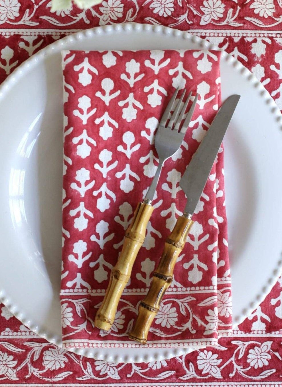 A styled table setting featuring red and white patterned placemats, a white plate, a matching folded napkin, gold-handled cutlery, and white flowers on a white wooden surface.