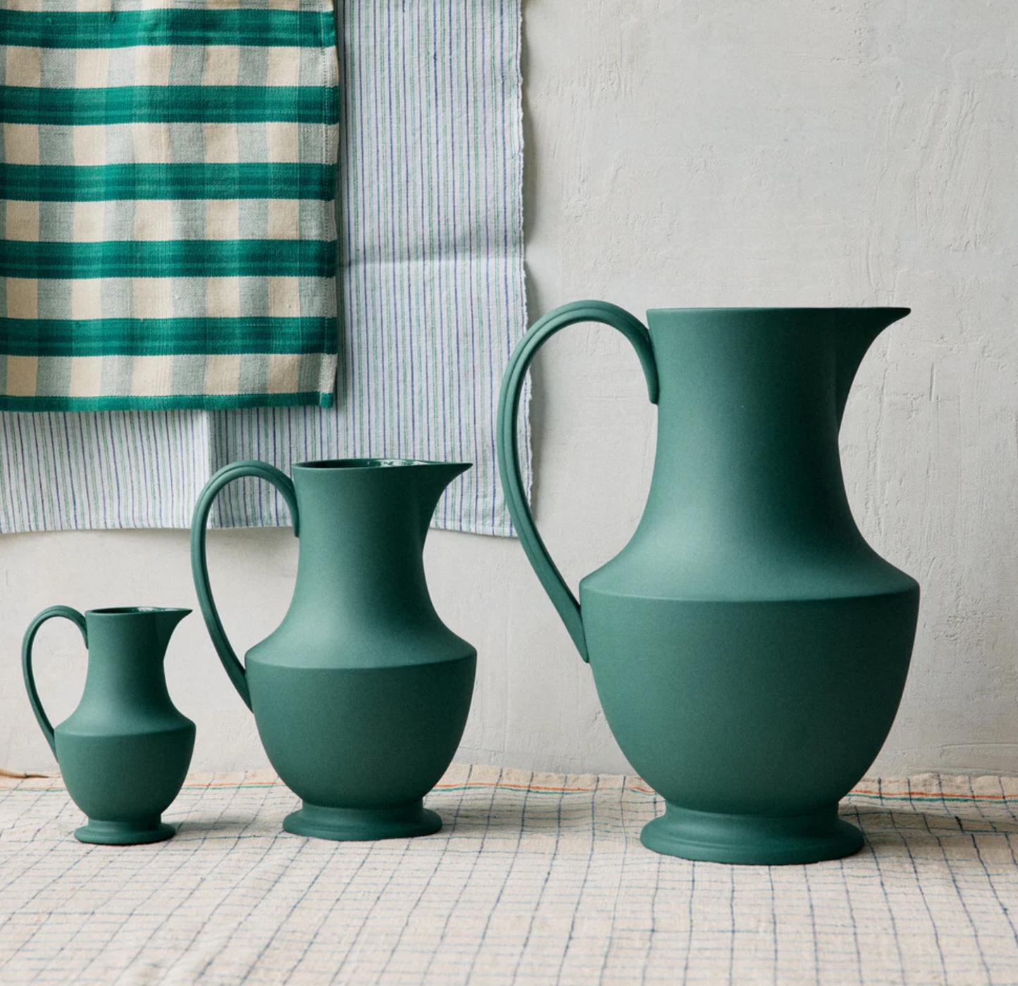 Three green ceramic jugs of different sizes on a checkered surface with a striped fabric background.