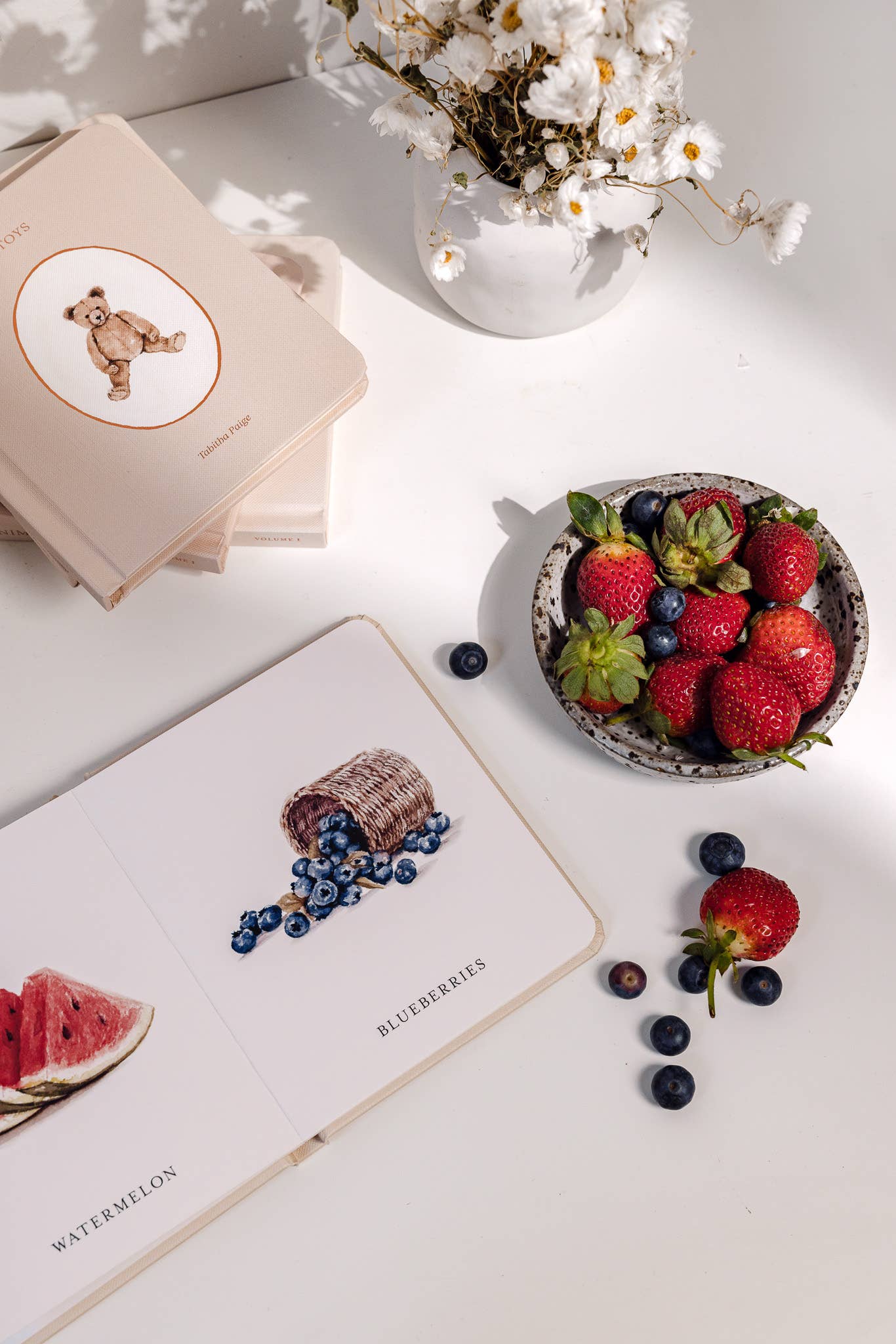 Bowl of strawberries and blueberries on a white surface with books and flowers.