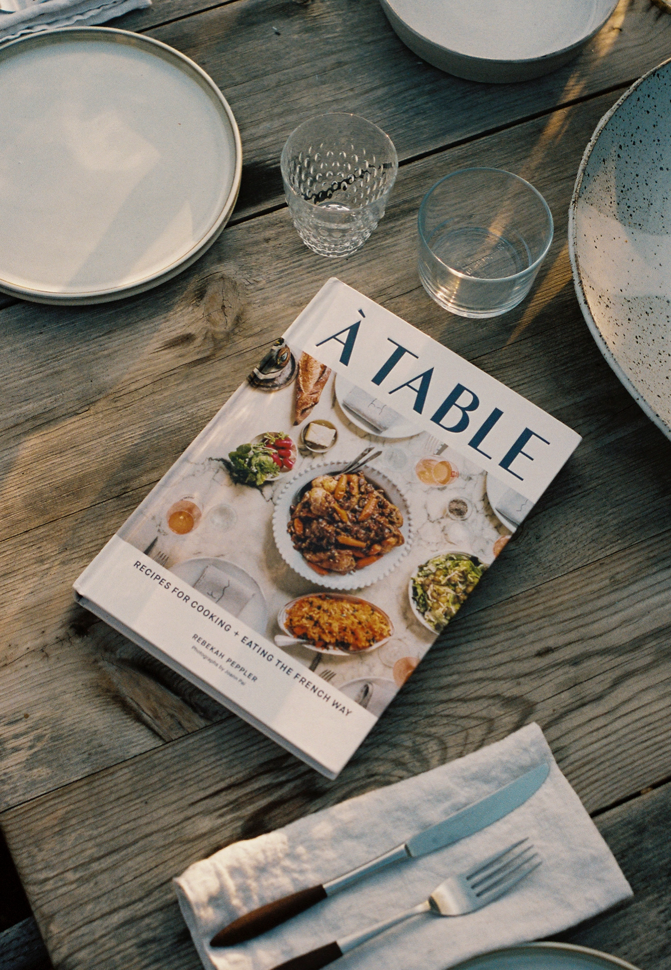 Cookbook titled 'A Table' on a wooden table with plates and glasses. Le Marchande Falmouth Maine 