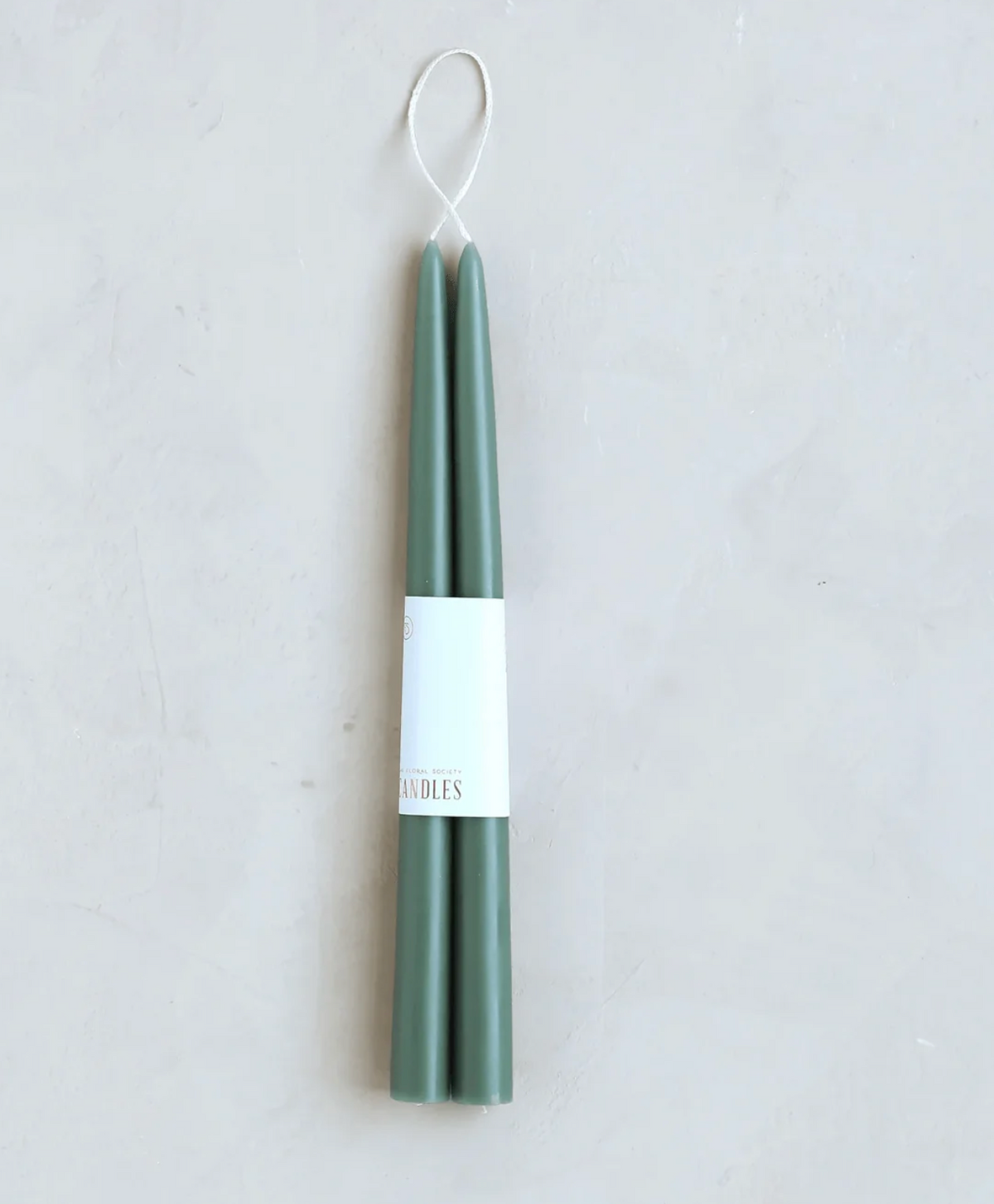 A set of two tall sage green taper candles in a white paper sleeve, hanging from a white loop string against a light gray textured background.