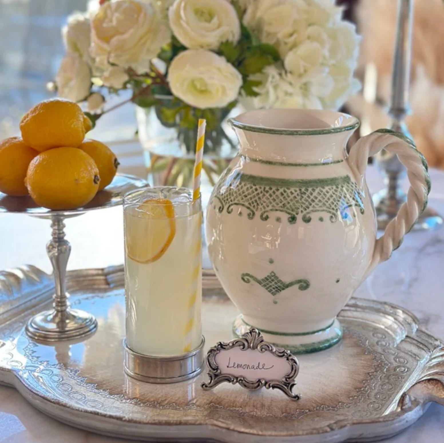Decorative table setting with a pitcher, glass of lemonade, and lemons on a silver tray.