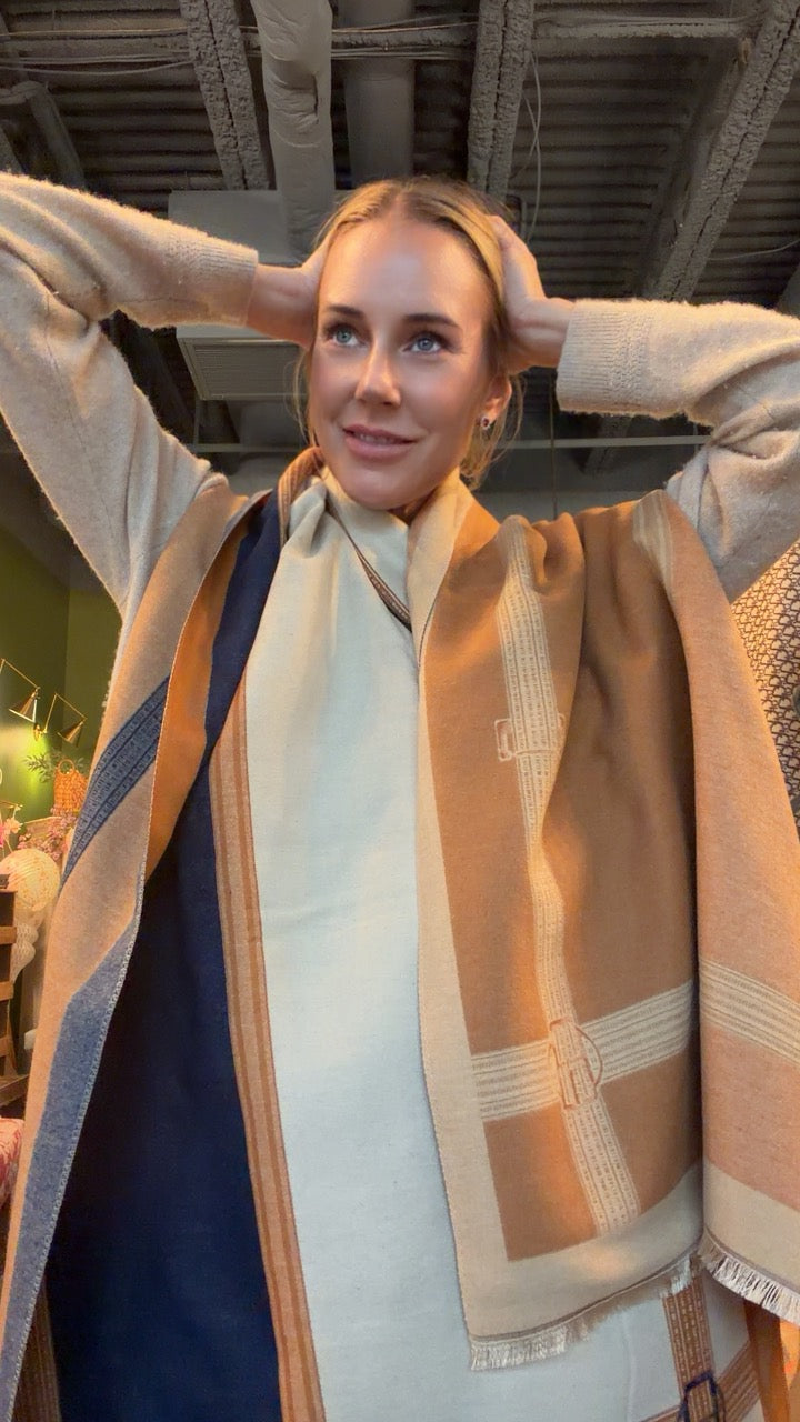A woman wearing a beige knit sweater and a large cream and tan plaid scarf with fringe, standing in a store with her hands behind her head.