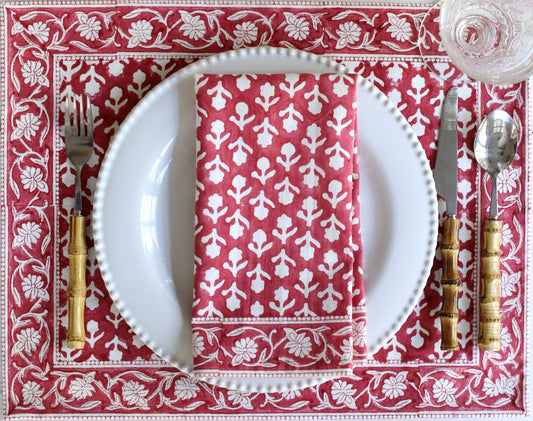 A styled table setting featuring a red and white patterned placemat with a matching folded napkin, white plates, gold-handled cutlery, and a glass of water.