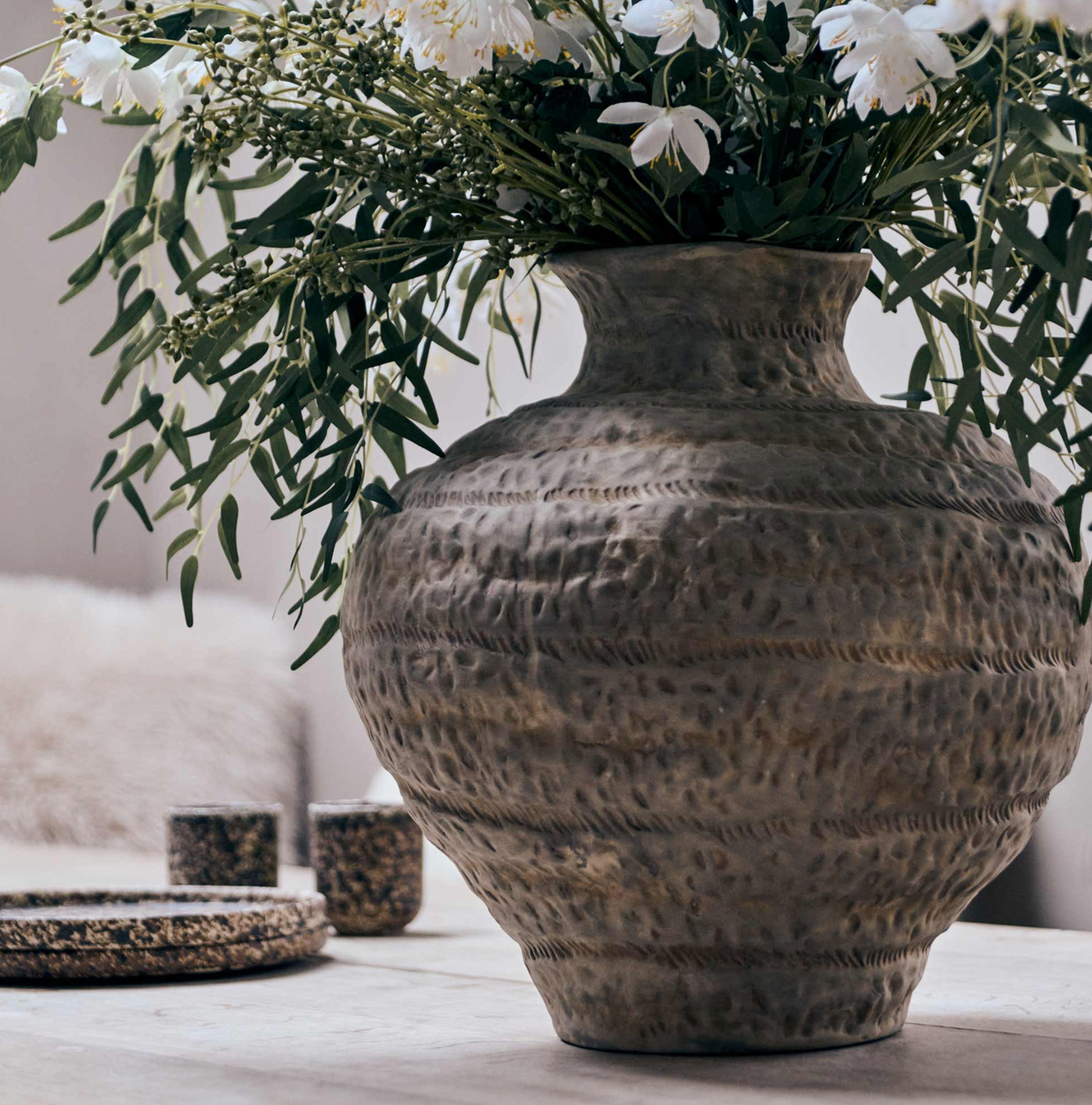 A large textured ceramic vase with a hammered finish, filled with white flowers and green foliage, set on a light stone surface in a softly lit interior.