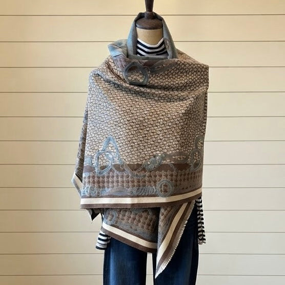 A patterned knit shawl draped over a mannequin torso, styled with a striped turtleneck and blue jeans against a white shiplap wall and wood floor.