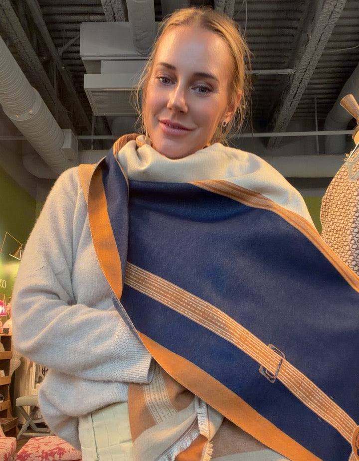 A woman wearing a light gray sweater and cream pants, wrapped in a large navy blue shawl with orange and cream striped borders, standing in a store interior.
