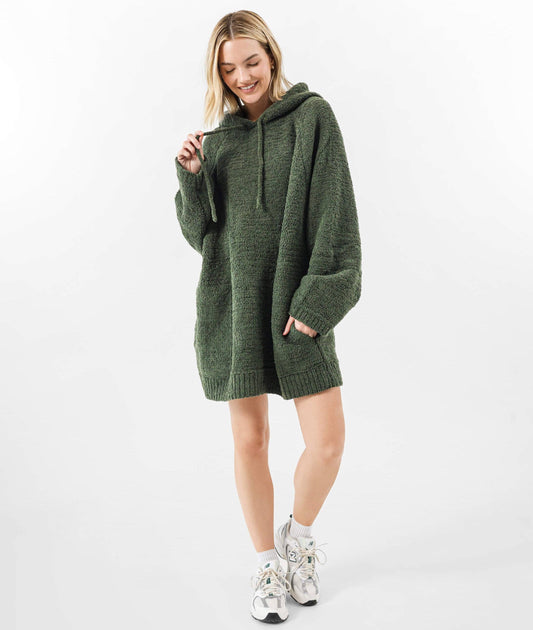 A woman modeling an oversized dark green knit hooded sweater dress with drawstrings and front pockets, paired with white socks and sneakers, against a plain white studio background.